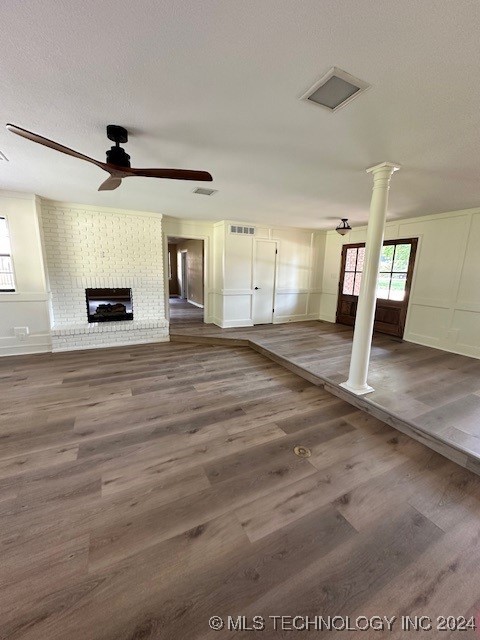 unfurnished living room with a textured ceiling, ornate columns, dark wood-type flooring, a brick fireplace, and ceiling fan