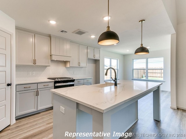 kitchen featuring stainless steel range with gas stovetop, light stone countertops, pendant lighting, a kitchen island with sink, and sink