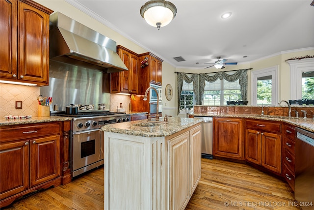 kitchen with light hardwood / wood-style flooring, appliances with stainless steel finishes, wall chimney exhaust hood, ceiling fan, and a kitchen island with sink