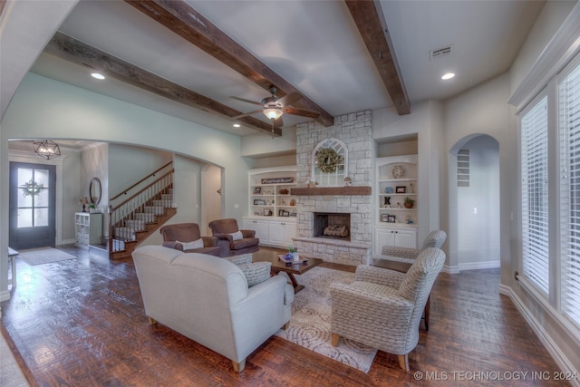 living room with a stone fireplace, ceiling fan, built in features, beam ceiling, and dark hardwood / wood-style flooring