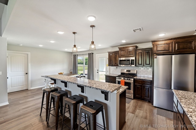 kitchen with appliances with stainless steel finishes, light hardwood / wood-style floors, dark brown cabinets, decorative light fixtures, and a kitchen island with sink