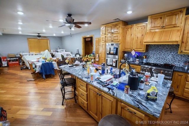 kitchen with light wood-type flooring, stainless steel appliances, a large island, dark stone counters, and ceiling fan