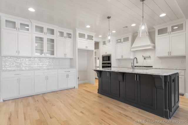 kitchen with custom exhaust hood, light wood-style flooring, stainless steel microwave, and white cabinets