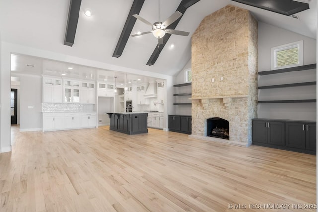 unfurnished living room with light wood-style flooring, ceiling fan, a stone fireplace, high vaulted ceiling, and beam ceiling