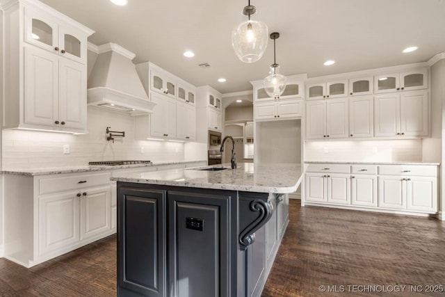 kitchen with premium range hood, a center island with sink, dark hardwood / wood-style flooring, light stone counters, and white cabinetry