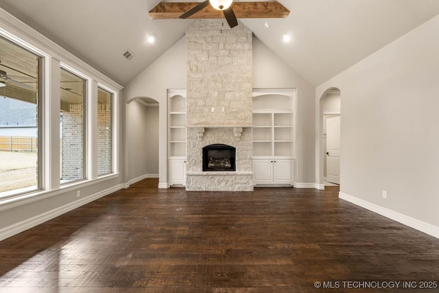 unfurnished living room with built in shelves, ceiling fan, dark wood-type flooring, beamed ceiling, and a stone fireplace