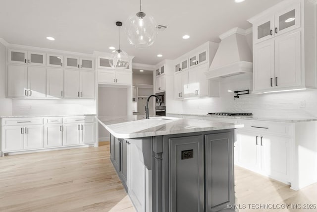 kitchen featuring premium range hood, light stone counters, a kitchen island with sink, pendant lighting, and white cabinets