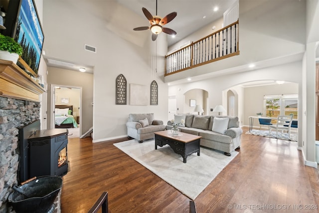 living room featuring a stone fireplace, ceiling fan, a towering ceiling, and hardwood / wood-style flooring