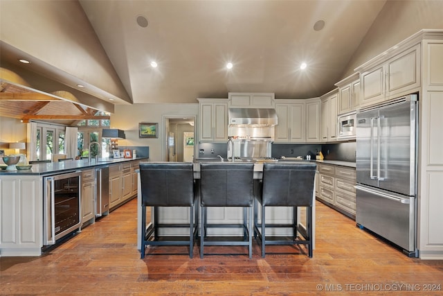 kitchen with light wood-type flooring, built in appliances, beverage cooler, vaulted ceiling, and a breakfast bar area