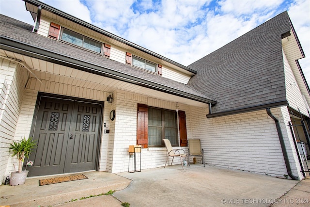 view of exterior entry with a porch, roof with shingles, and brick siding