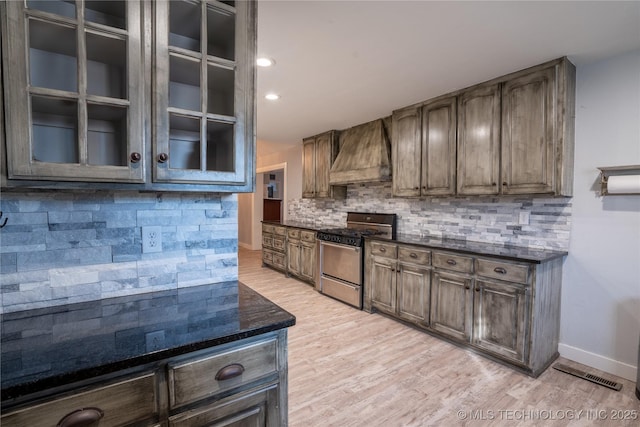 kitchen featuring custom exhaust hood, stainless steel gas range oven, backsplash, light wood-type flooring, and dark stone countertops