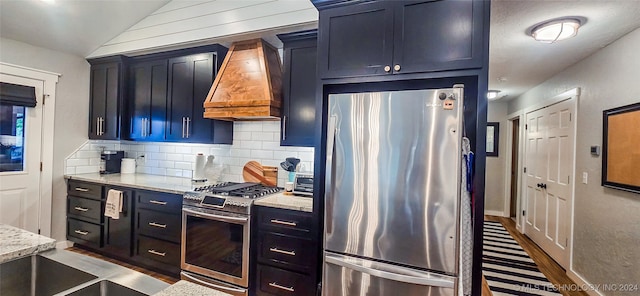 kitchen with appliances with stainless steel finishes, tasteful backsplash, light stone counters, custom range hood, and blue cabinets