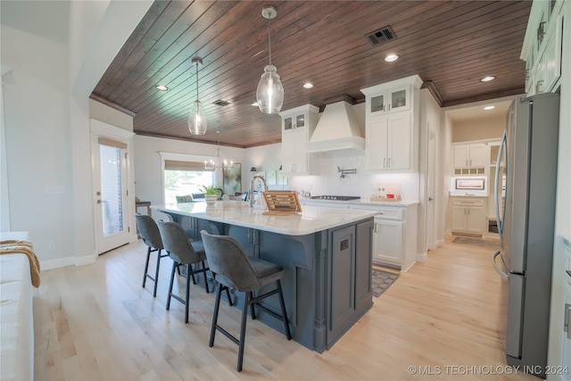 kitchen featuring wood ceiling, white cabinetry, custom exhaust hood, a center island with sink, and stainless steel fridge