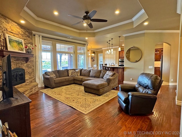 living room featuring ornamental molding, a tray ceiling, a fireplace, ceiling fan with notable chandelier, and dark hardwood / wood-style flooring