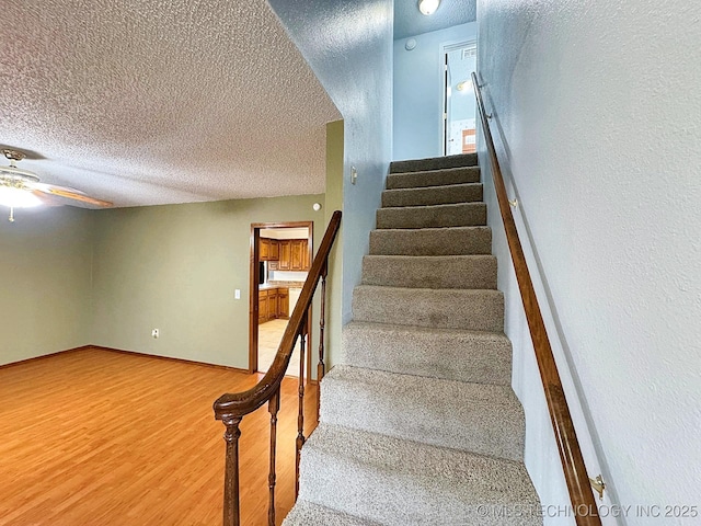 stairway with a textured ceiling, wood finished floors, a ceiling fan, and baseboards