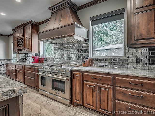 kitchen featuring dark brown cabinetry, premium range hood, range with two ovens, crown molding, and decorative backsplash