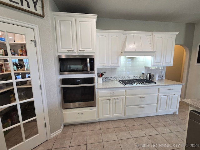 kitchen with white cabinets, light tile patterned floors, appliances with stainless steel finishes, custom range hood, and decorative backsplash