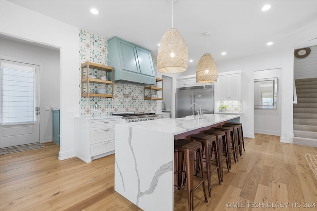 kitchen featuring stainless steel built in refrigerator, an island with sink, white cabinets, hanging light fixtures, and light hardwood / wood-style flooring