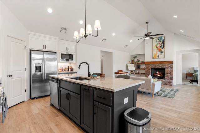 kitchen with sink, ceiling fan with notable chandelier, a brick fireplace, a center island with sink, and appliances with stainless steel finishes