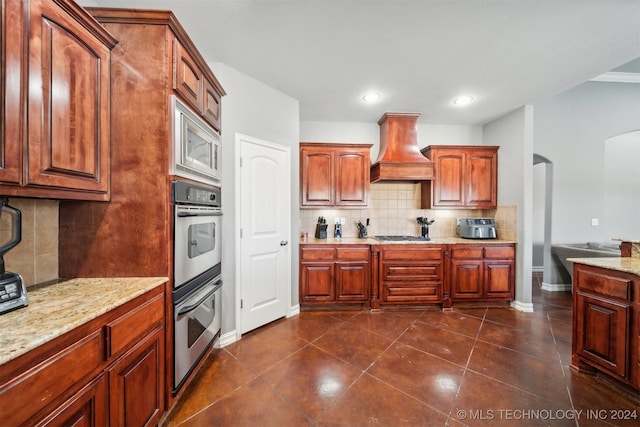 kitchen with custom exhaust hood, dark tile patterned flooring, backsplash, appliances with stainless steel finishes, and light stone countertops