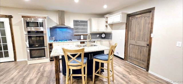 kitchen with white cabinets, an island with sink, wall chimney range hood, stainless steel appliances, and light stone countertops