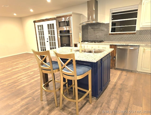 kitchen with appliances with stainless steel finishes, light hardwood / wood-style floors, light stone countertops, a center island with sink, and wall chimney range hood