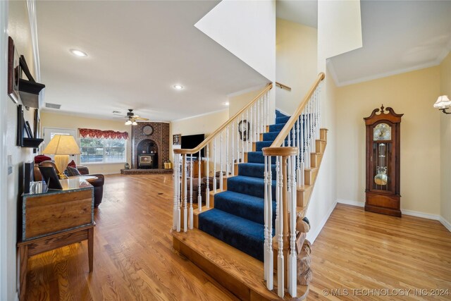 stairs with wood-type flooring, ornamental molding, ceiling fan, and a brick fireplace