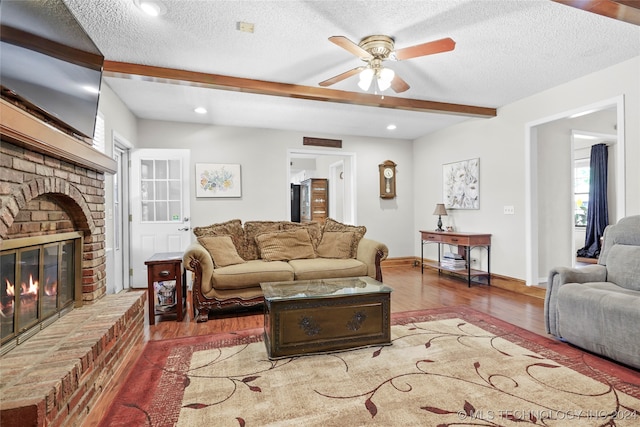 living room with a brick fireplace, a textured ceiling, ceiling fan, beamed ceiling, and hardwood / wood-style floors