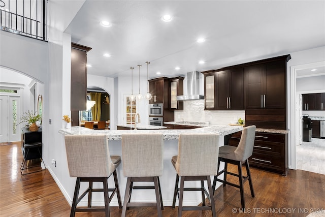 kitchen featuring decorative light fixtures, wall chimney exhaust hood, dark wood-type flooring, appliances with stainless steel finishes, and a kitchen bar