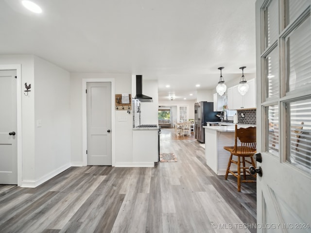 kitchen featuring stainless steel fridge, hanging light fixtures, white cabinets, wall chimney exhaust hood, and light wood-type flooring