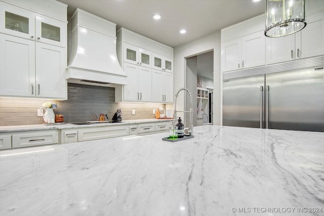 kitchen featuring stainless steel built in refrigerator, white cabinets, light stone countertops, and custom range hood