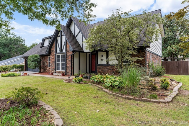 tudor-style house featuring brick siding, roof with shingles, stucco siding, fence, and a front lawn