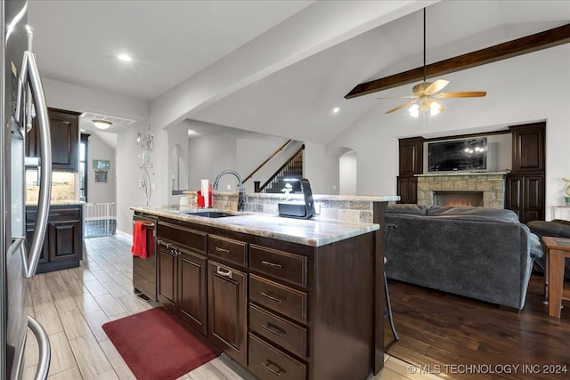 kitchen with ceiling fan, an island with sink, sink, a stone fireplace, and lofted ceiling with beams