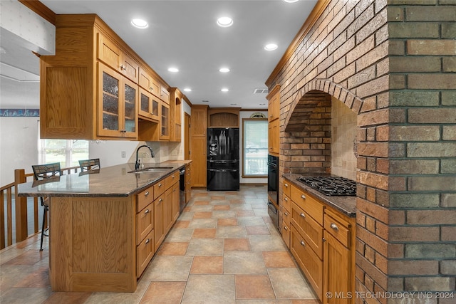 kitchen featuring glass insert cabinets, black appliances, a kitchen bar, dark stone counters, and a sink