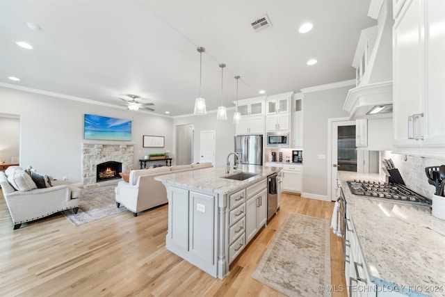 kitchen with appliances with stainless steel finishes, white cabinets, a stone fireplace, ceiling fan, and a center island with sink