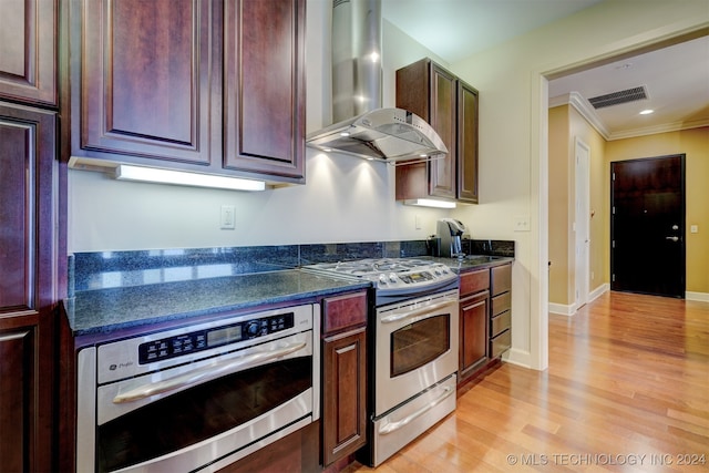 kitchen featuring ornamental molding, wall chimney exhaust hood, stainless steel appliances, dark stone countertops, and light wood-type flooring