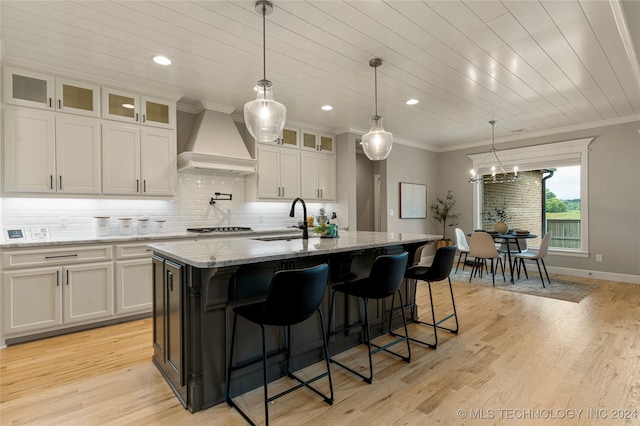 kitchen with light stone counters, an island with sink, white cabinets, premium range hood, and sink