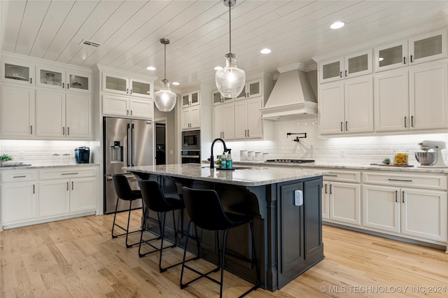 kitchen featuring light stone counters, custom exhaust hood, an island with sink, white cabinetry, and stainless steel appliances