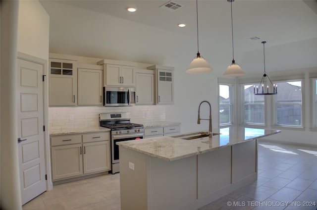 kitchen featuring appliances with stainless steel finishes, an island with sink, light stone counters, decorative light fixtures, and sink