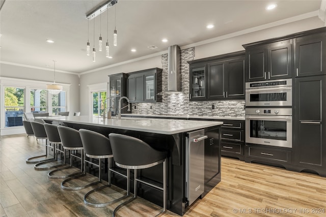 kitchen featuring double oven, a large island with sink, pendant lighting, and wall chimney range hood