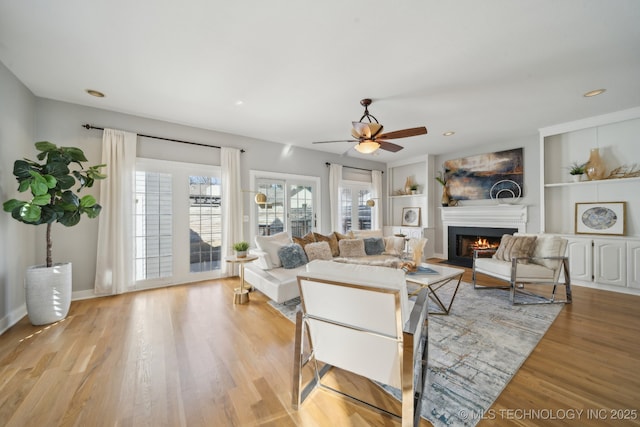 living area featuring built in shelves, a ceiling fan, wood finished floors, recessed lighting, and a lit fireplace