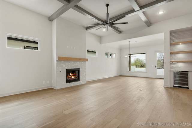 unfurnished living room with light wood-type flooring, wine cooler, beamed ceiling, ceiling fan with notable chandelier, and a fireplace