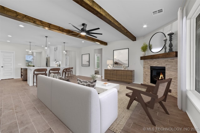 living room with ceiling fan, a fireplace, sink, and beamed ceiling