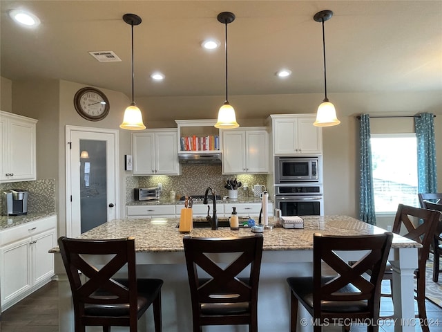 kitchen with light stone counters, a kitchen island with sink, white cabinetry, appliances with stainless steel finishes, and decorative light fixtures