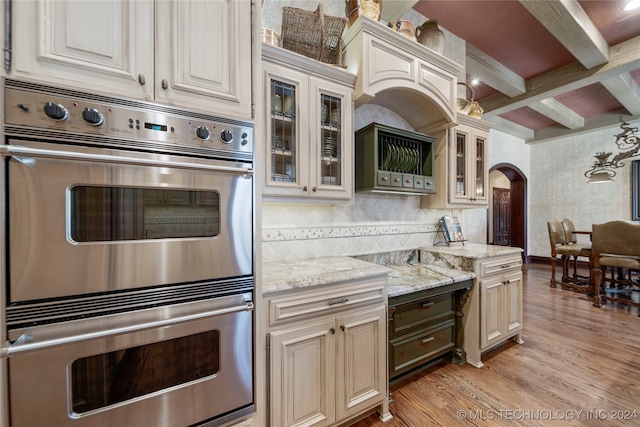 kitchen with light stone counters, beam ceiling, backsplash, double oven, and light hardwood / wood-style floors