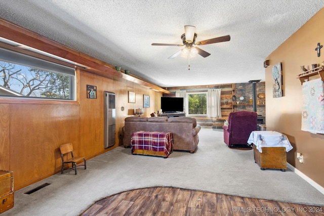 carpeted living room with ceiling fan, a wood stove, a textured ceiling, and wooden walls