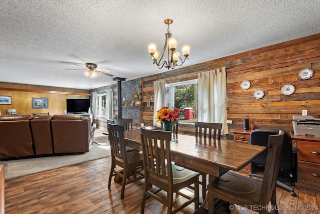 dining room featuring wood walls, a textured ceiling, a wood stove, hardwood / wood-style floors, and ceiling fan with notable chandelier