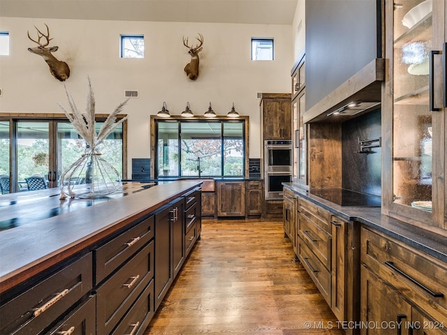 kitchen with dark brown cabinetry, stainless steel double oven, wall chimney range hood, black electric stovetop, and light wood-type flooring