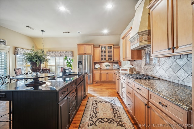 kitchen with appliances with stainless steel finishes, premium range hood, light wood-type flooring, a center island with sink, and sink