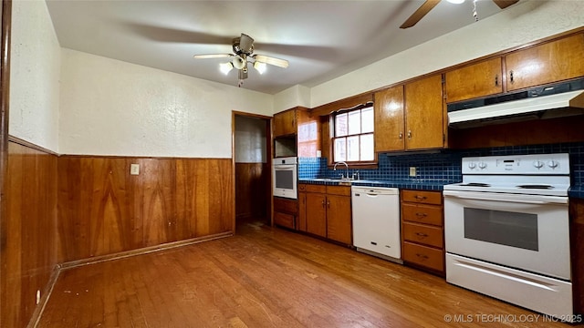 kitchen featuring wooden walls, under cabinet range hood, white appliances, wainscoting, and brown cabinetry